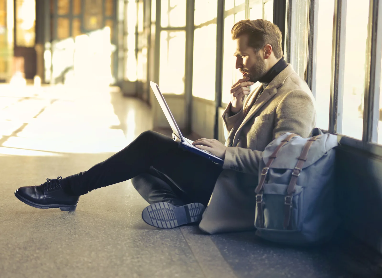 Mann sitter på gulvet i en flyplassterminal med laptop på fanget og ryggsekken ved siden av seg