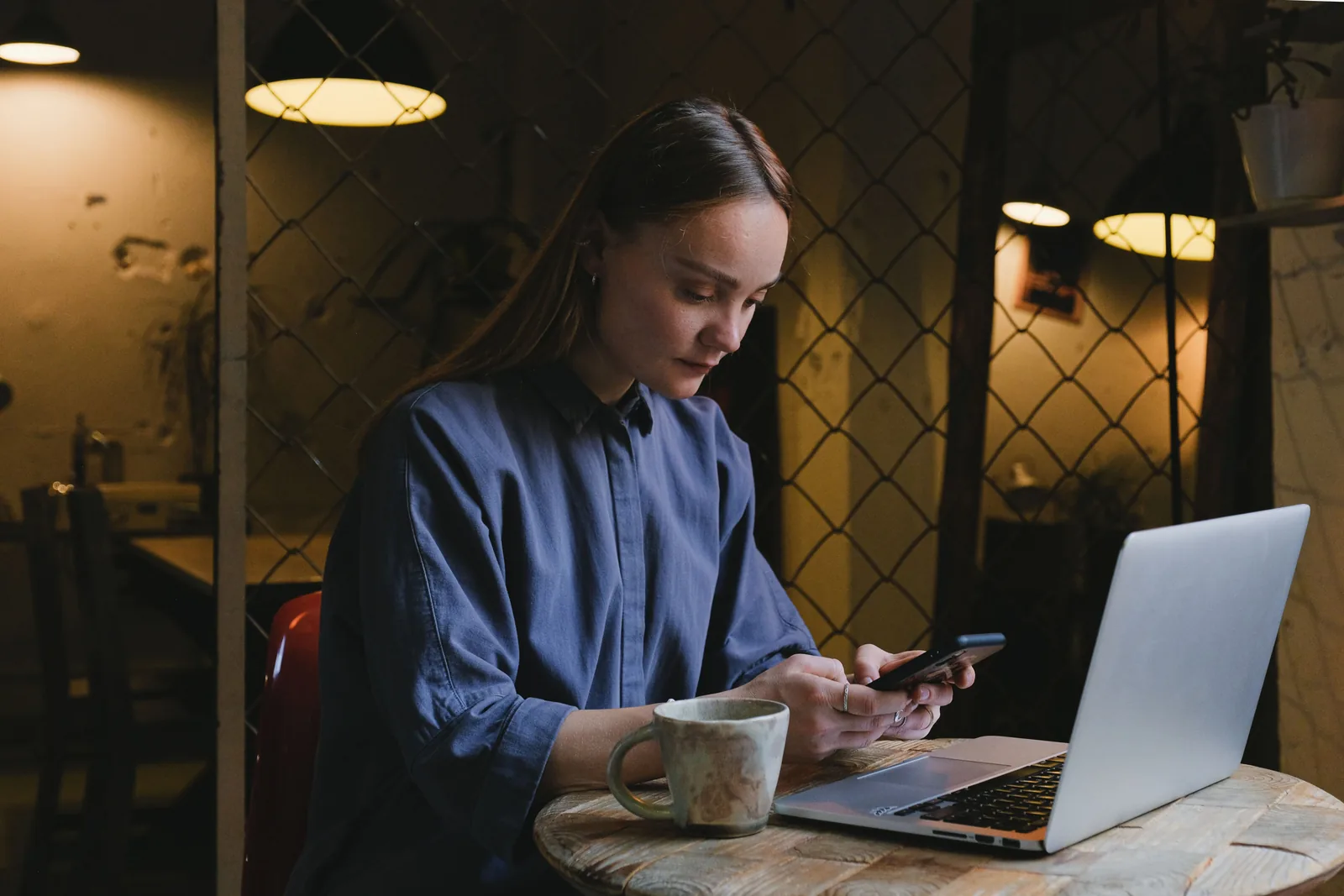 Kvinne på kafé med kaffekopp, mobil i hånden og laptop på bordet