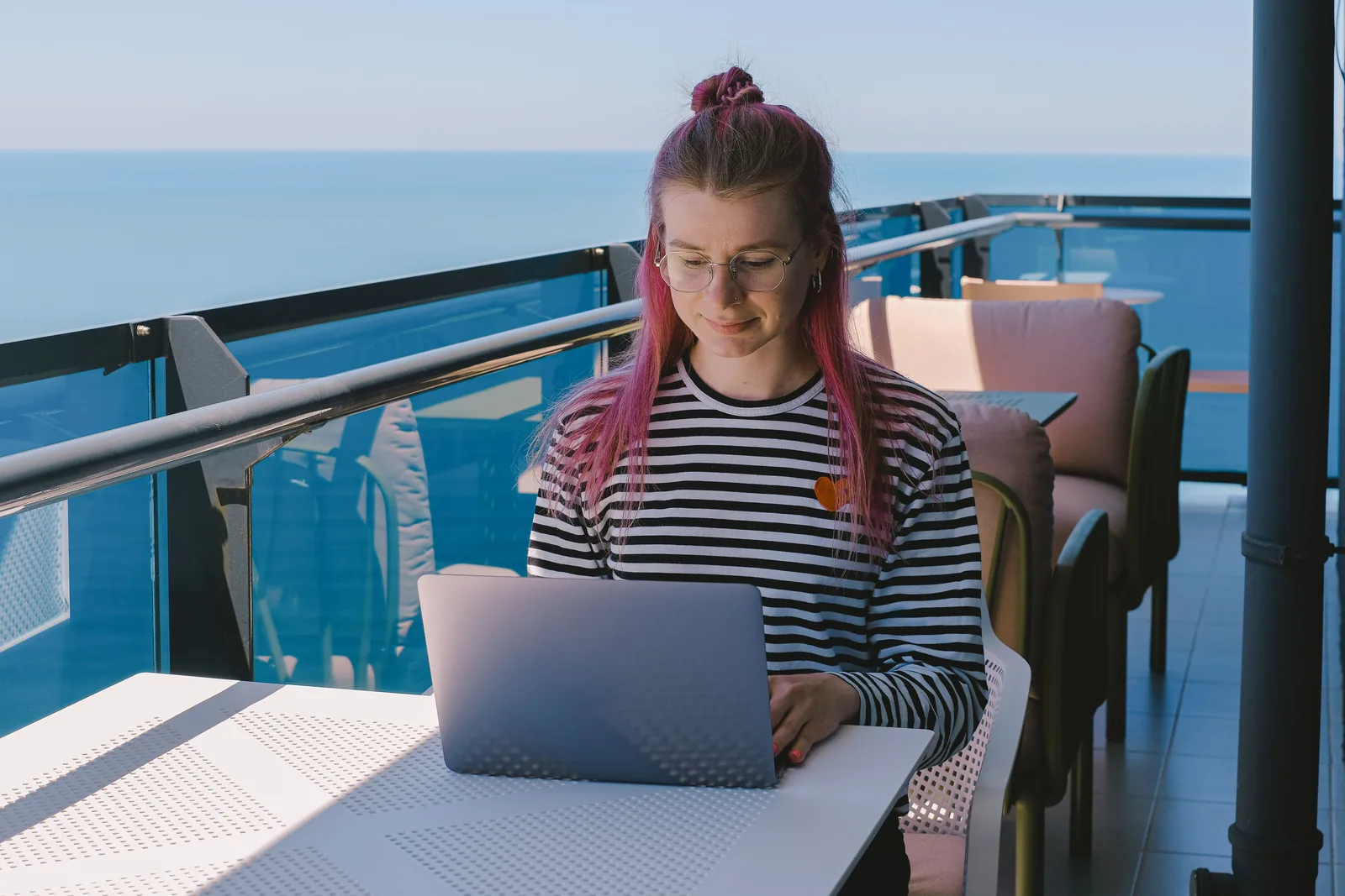 Kvinne i stripete genser jobber på laptop ved et bord på en hotellbalkong med utsikt over havet
