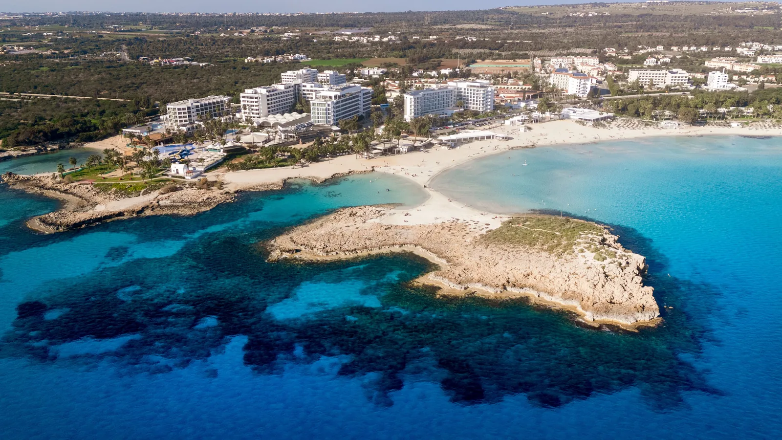 Flyfoto av en turkis bukt og en liten holme ved Ayia Napa på Kypros, med hoteller langs kysten