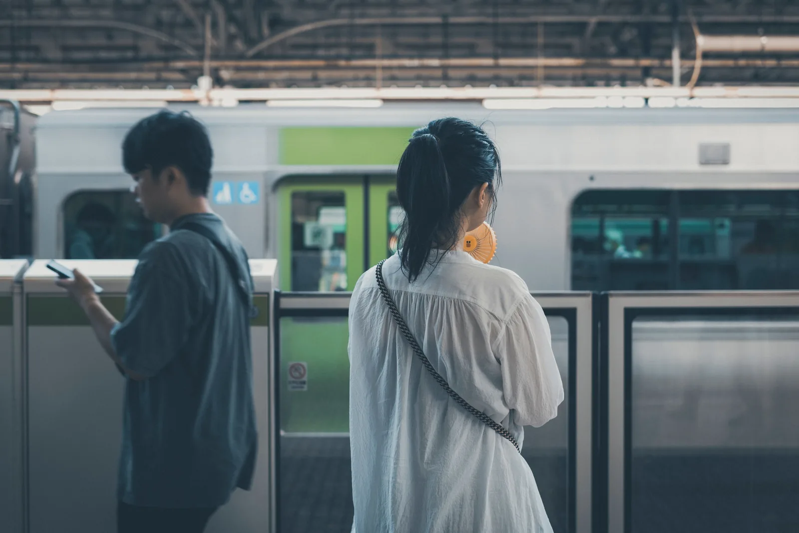 To reisende venter bak plattformdørene på en togstasjon i Tokyo, mannen til venstre ser på mobilen