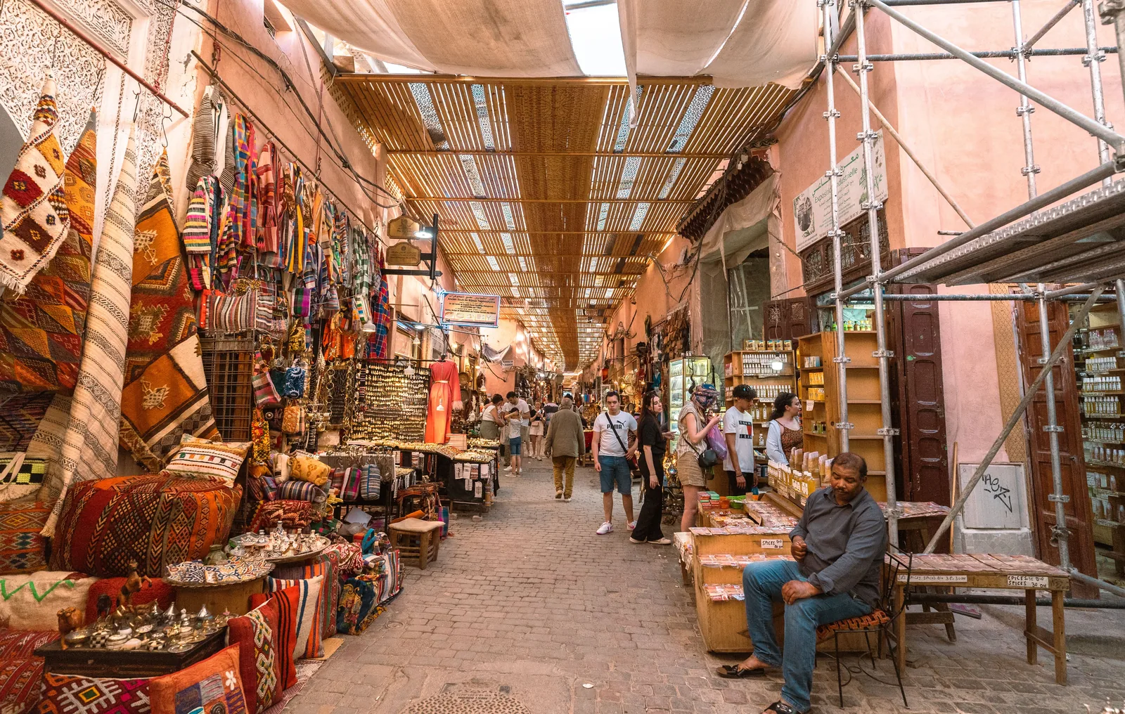 Smal souk-gate i Marrakech med tepper, tekstiler og lamper utenfor butikkene, og folk som går i det overbygde markedet