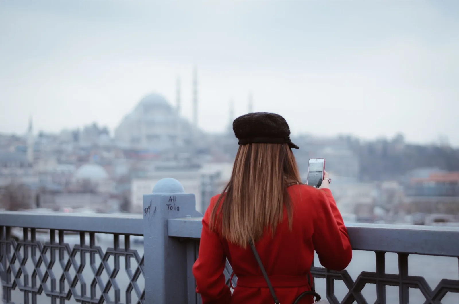 Kvinne i rød kåpe fotograferer utsikten over Istanbul med mobilen fra en bro, med en stor moské i bakgrunnen