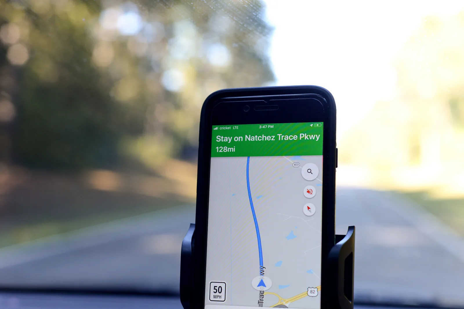 Mobiltelefon i holder på dashbordet viser kartnavigasjon langs Natchez Trace Parkway, med landeveien uskarp i bakgrunnen