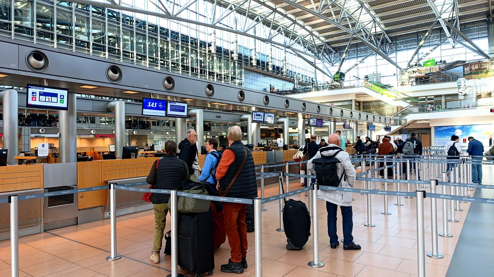 Reisende står i kø foran innsjekkingsskranker i en lys terminalhall på Hamburg lufthavn