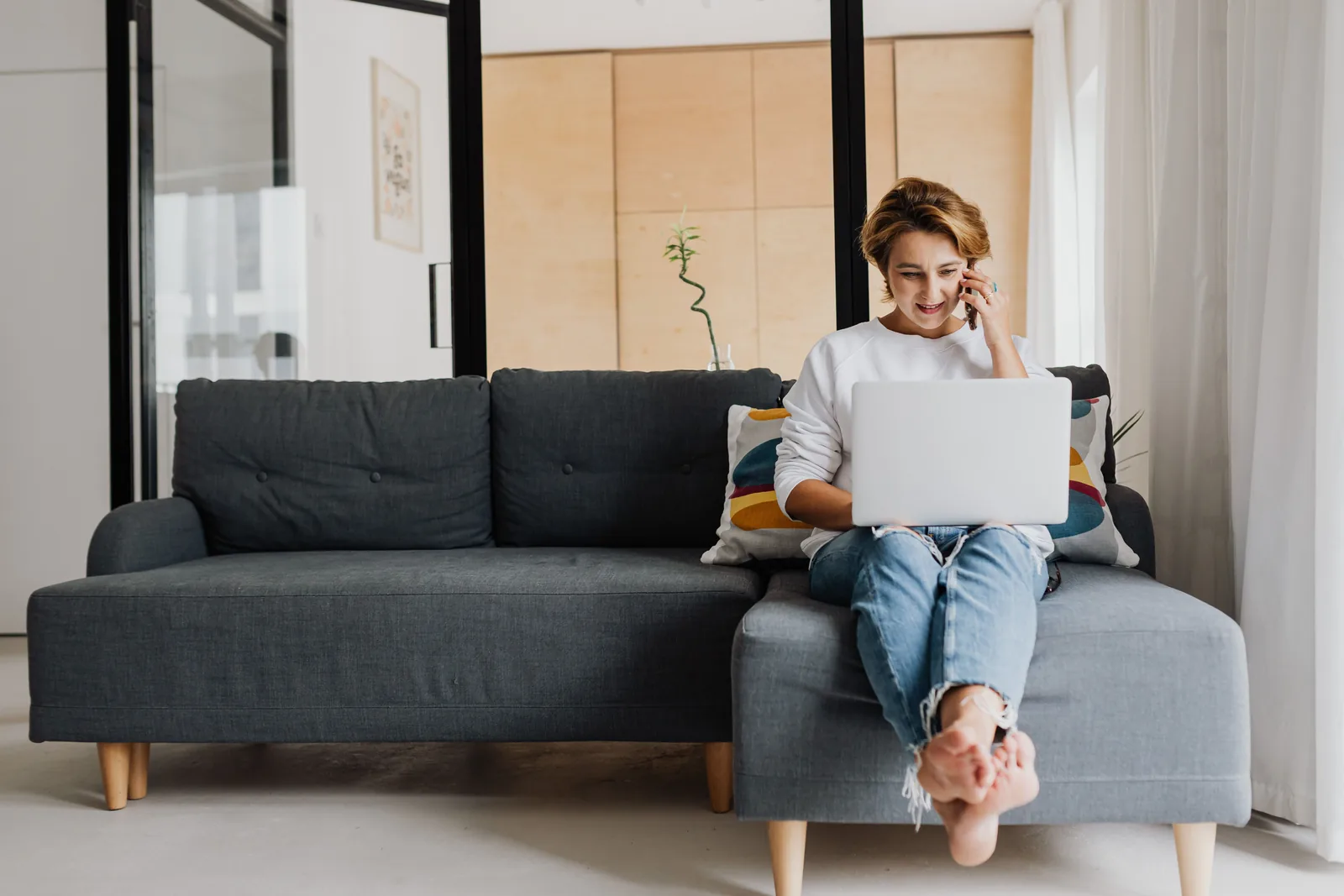 Kvinne sitter i en grå sofa med laptop på fanget og snakker i mobiltelefonen