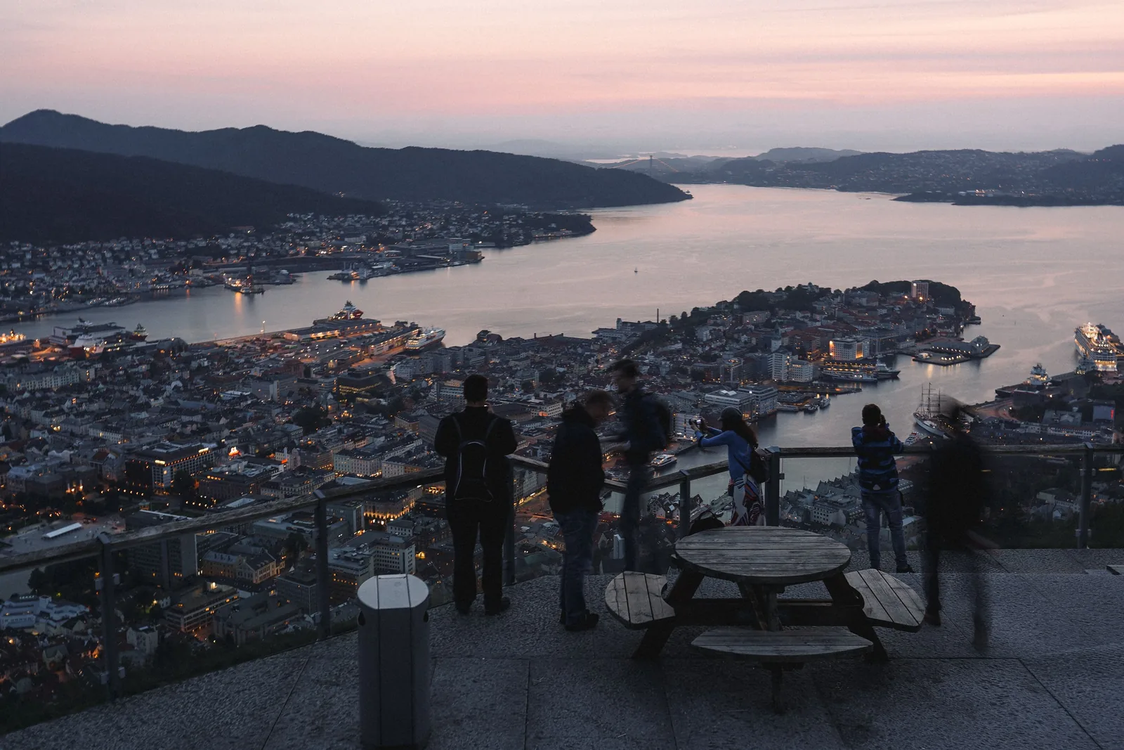 Utsikt over Bergen sentrum og havna i skumringen fra et utsiktspunkt i fjellet, med skip ved kai og folk ved rekkverket