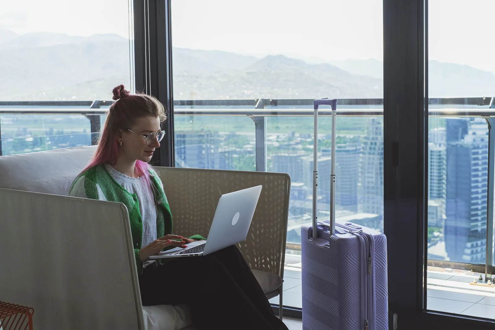 Kvinne med laptop i en hotellounge, med en lilla koffert ved siden av og utsikt over byen gjennom vinduet