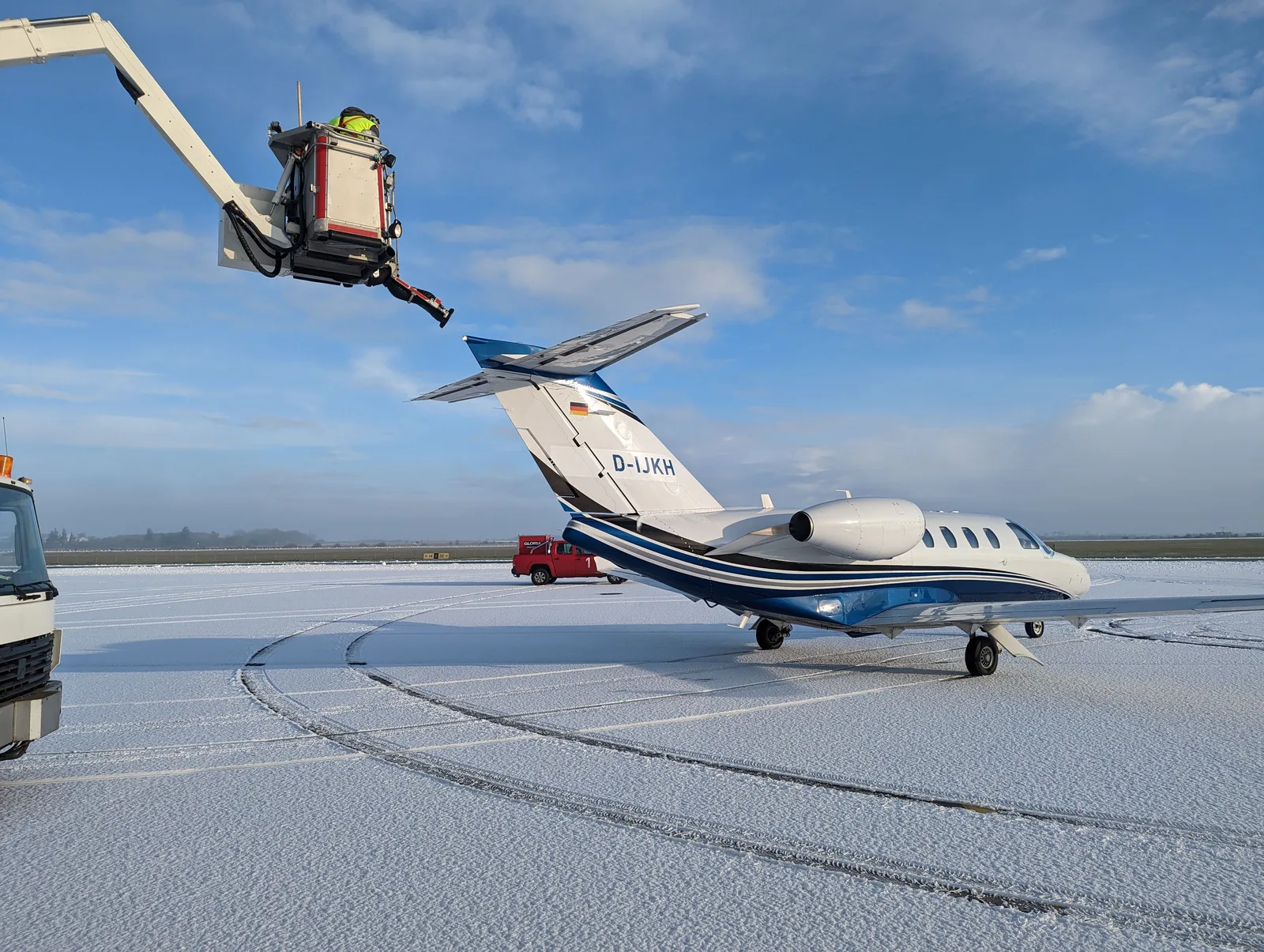 Avising av et lite fly på en snødekt flyplass en solrik vinterdag, med en arbeider i lift ved halen
