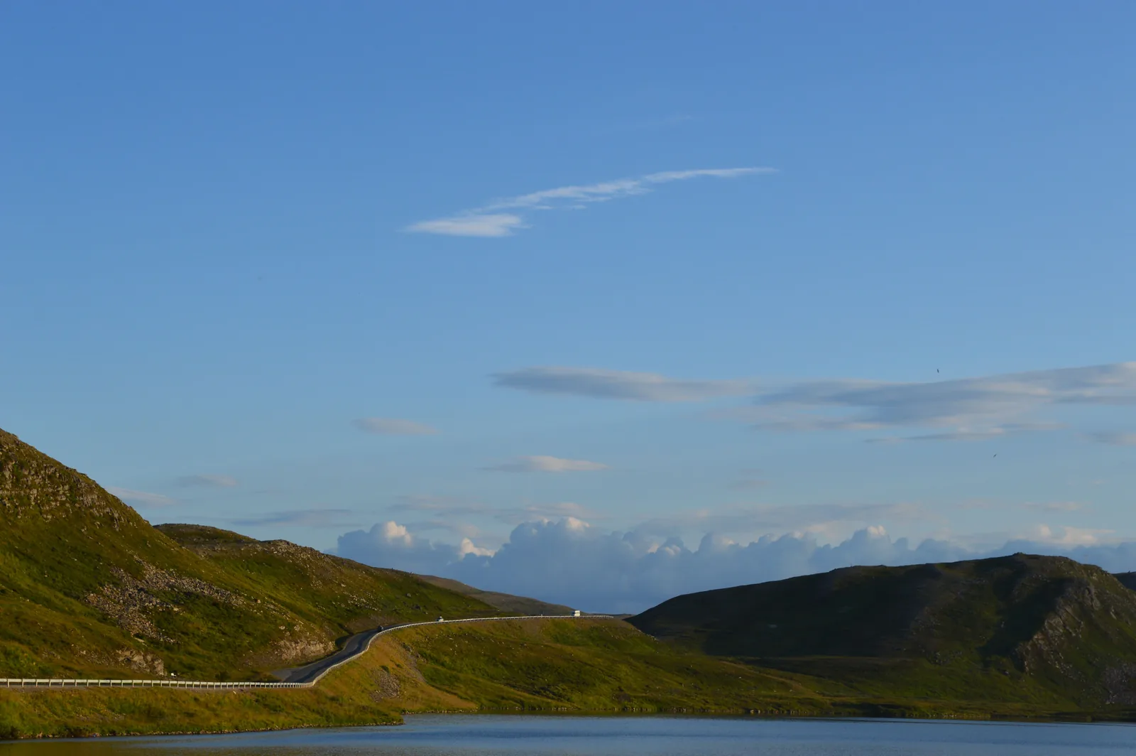 Vei med autovern som slynger seg langs en grønn åsside og et vann i Finnmark en sommerkveld