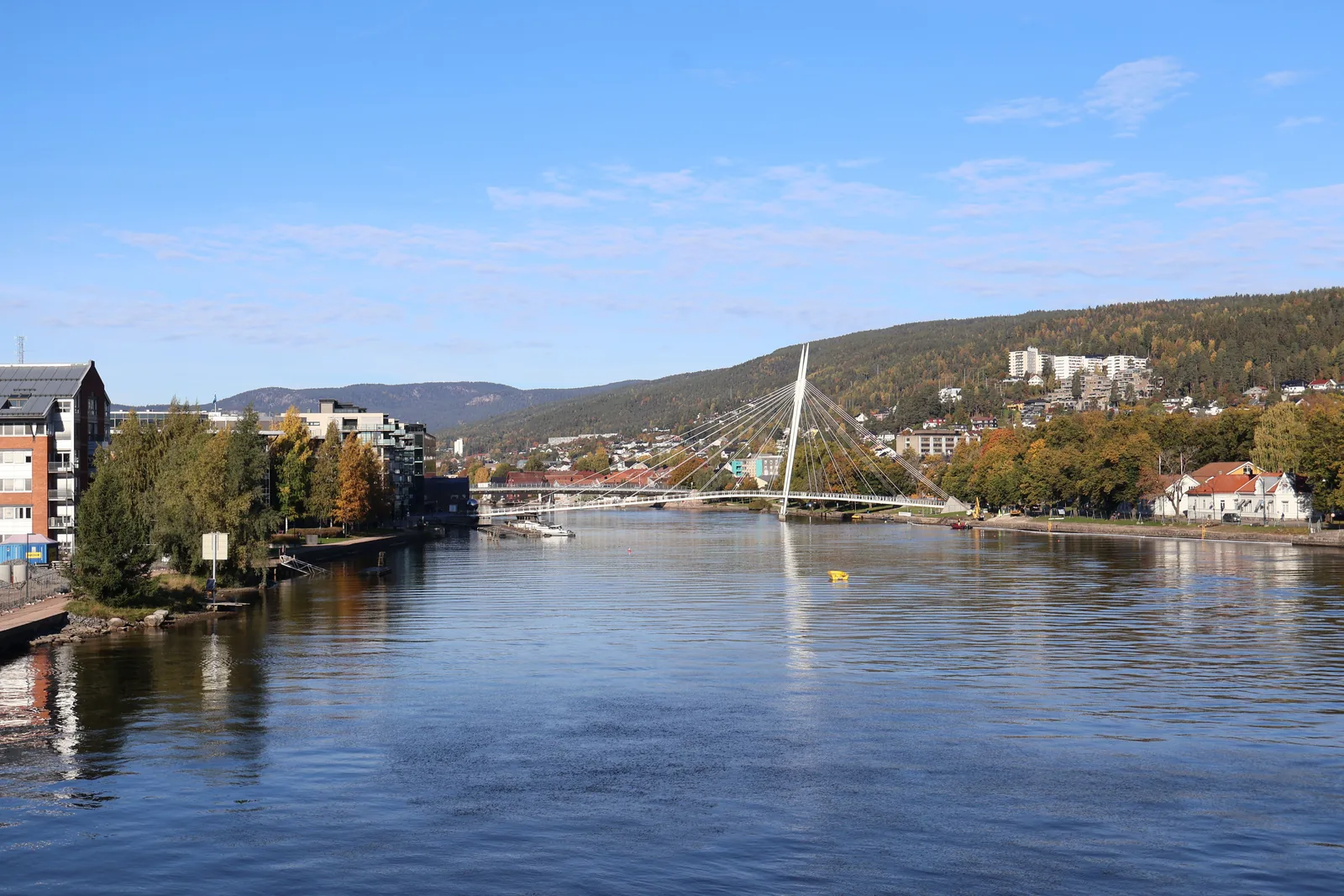 Gangbru med skråstag over Drammenselva en solrik høstdag, med bebyggelse og skogkledde åser i bakgrunnen