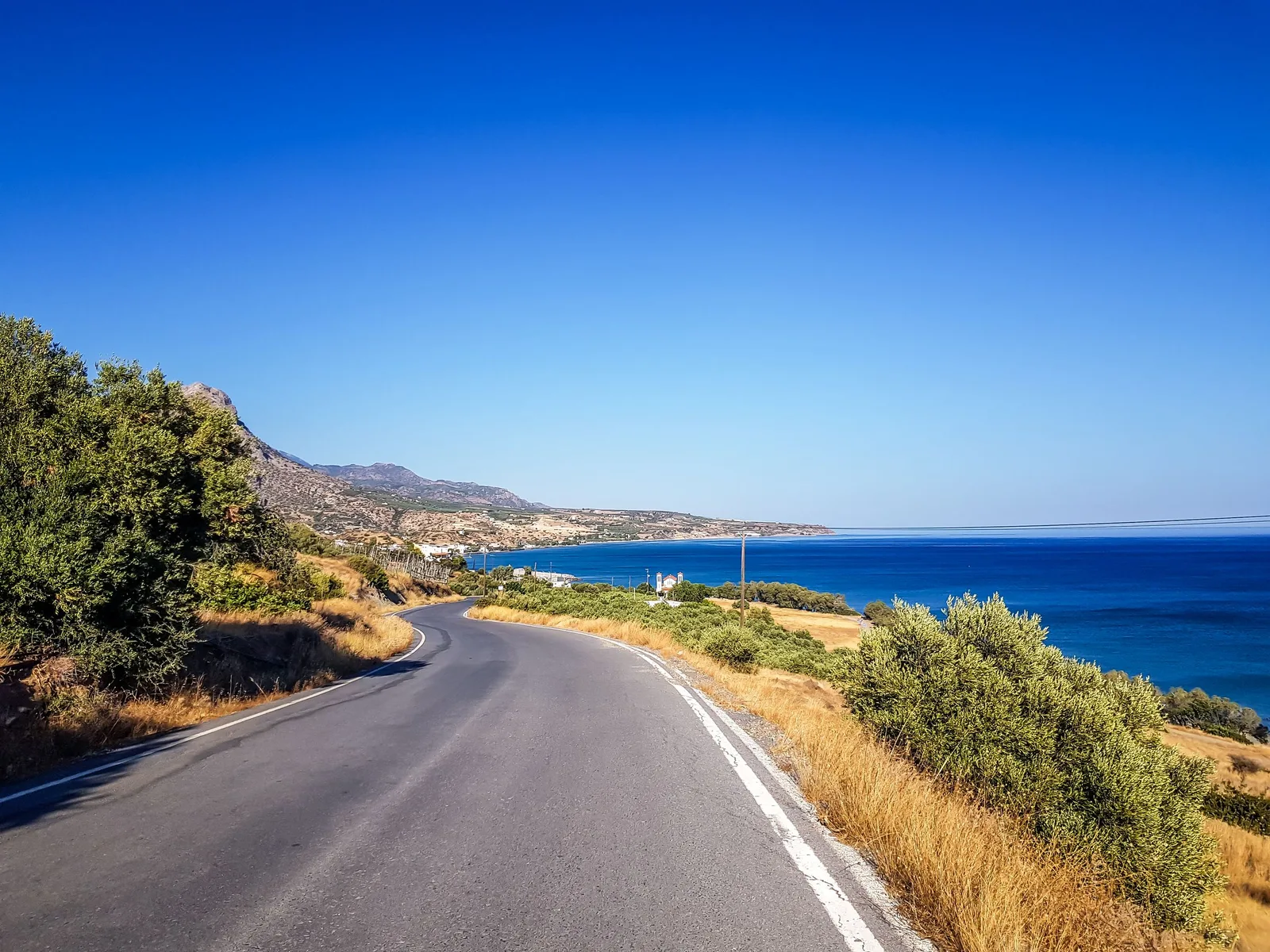 Smal asfaltert vei langs kysten på Kreta med blått hav og tørt kratt langs veikanten