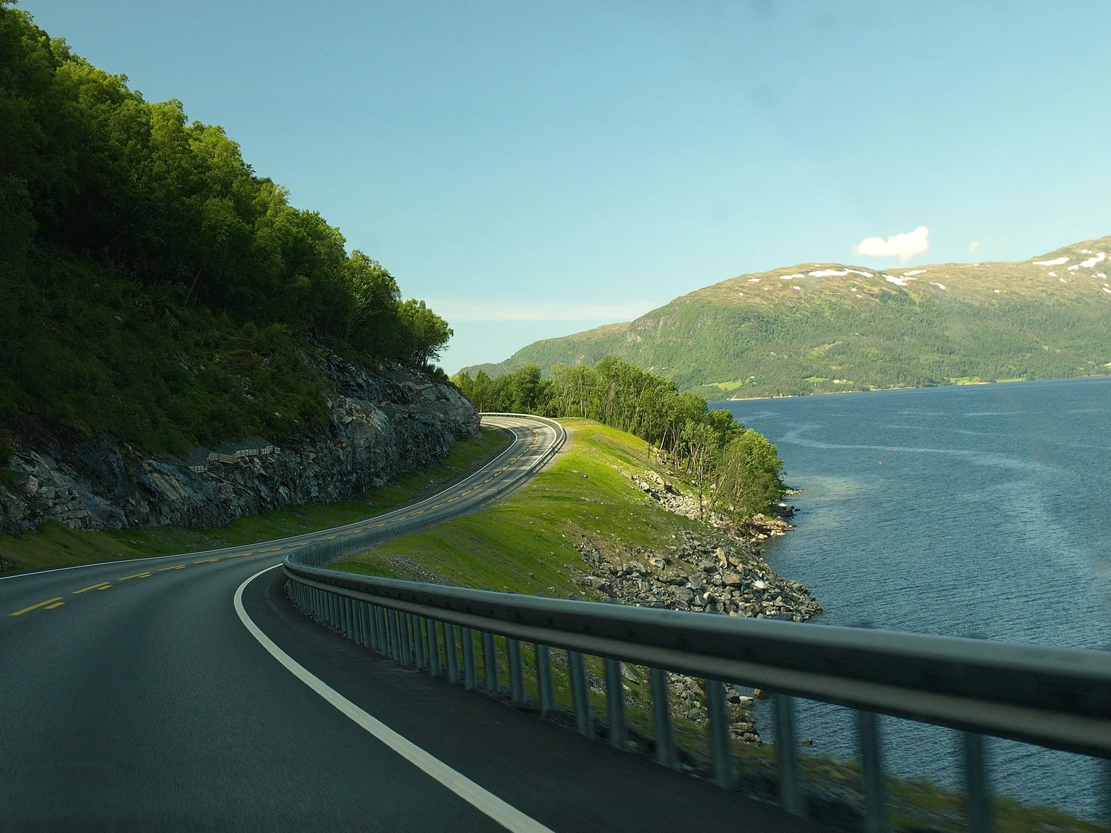 Landevei som svinger langs en fjord med grønne fjell og blå himmel på Vestlandet