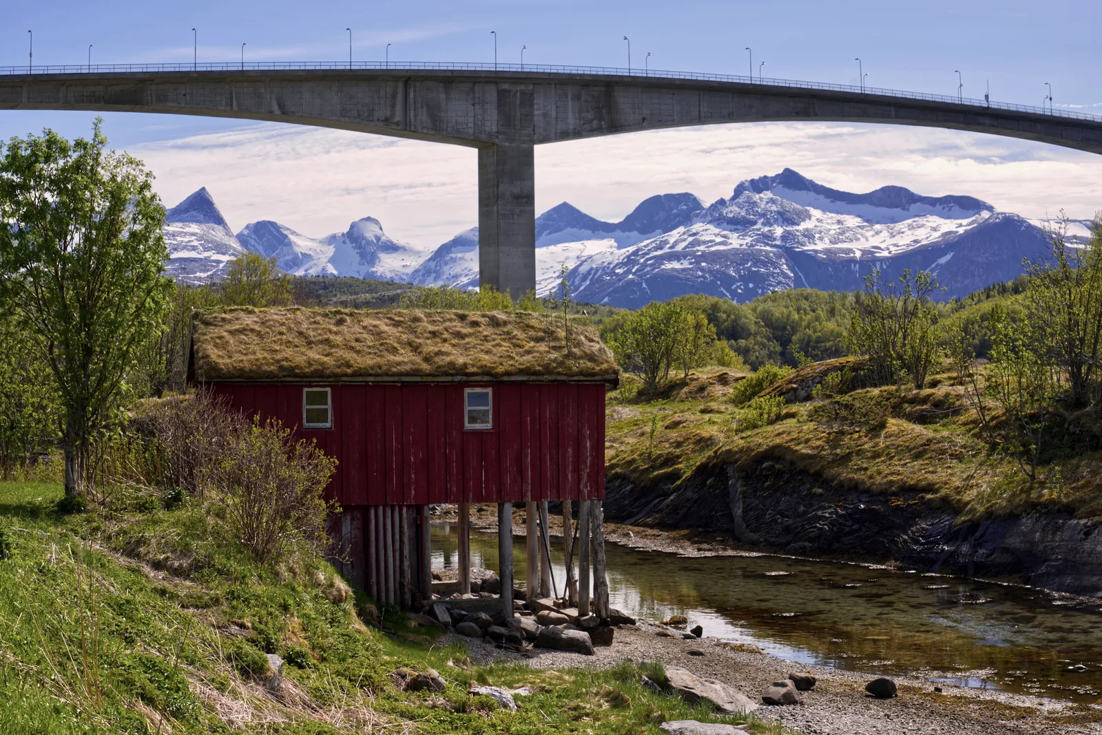 Saltstraumbrua og en rød sjøbod med torvtak foran snødekte fjell ved Saltstraumen