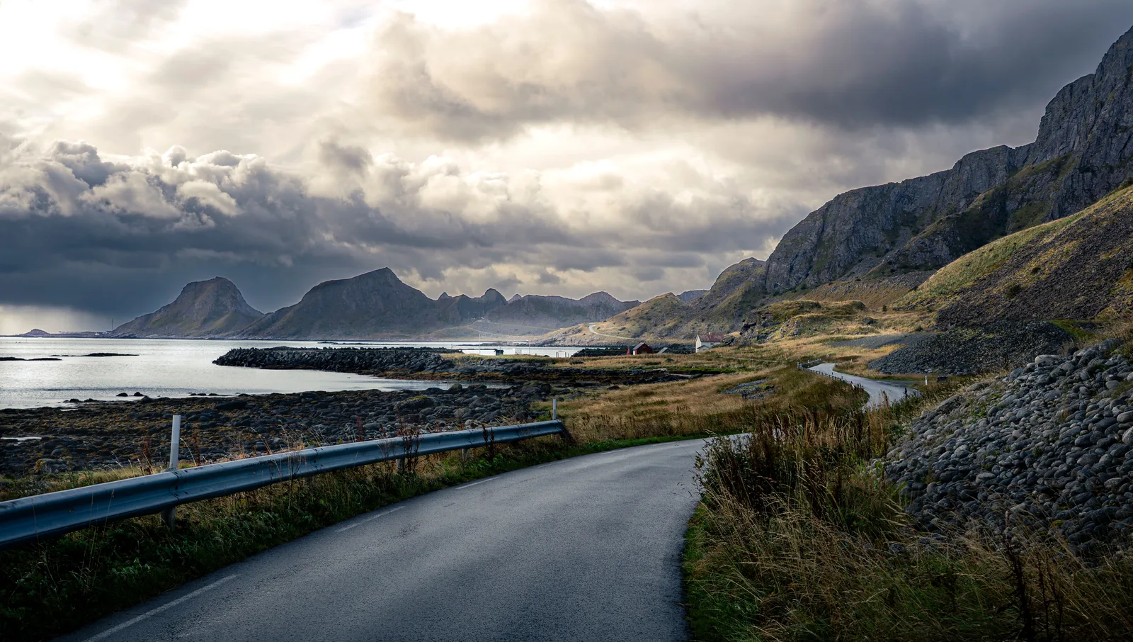 Smal asfaltvei langs kysten under mørke skyer, med bratte fjell og havet i bakgrunnen på Værøy i Lofoten