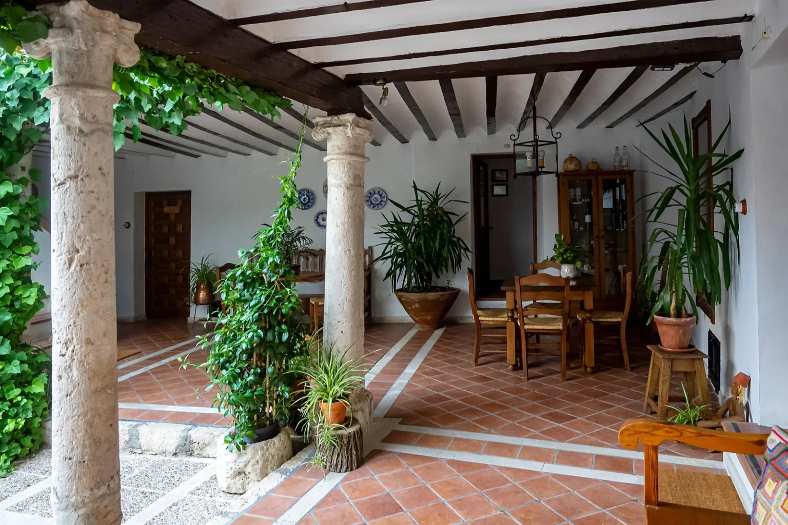 Patio i et gammelt spansk hus i Chinchón med trebjelker i taket, søyler, rødt flisgulv, et spisebord med stoler og mange grønne planter i krukker