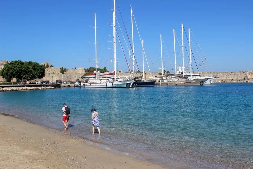 Sandstrand på Rhodos med folk i vannkanten og seilbåter ved en molo