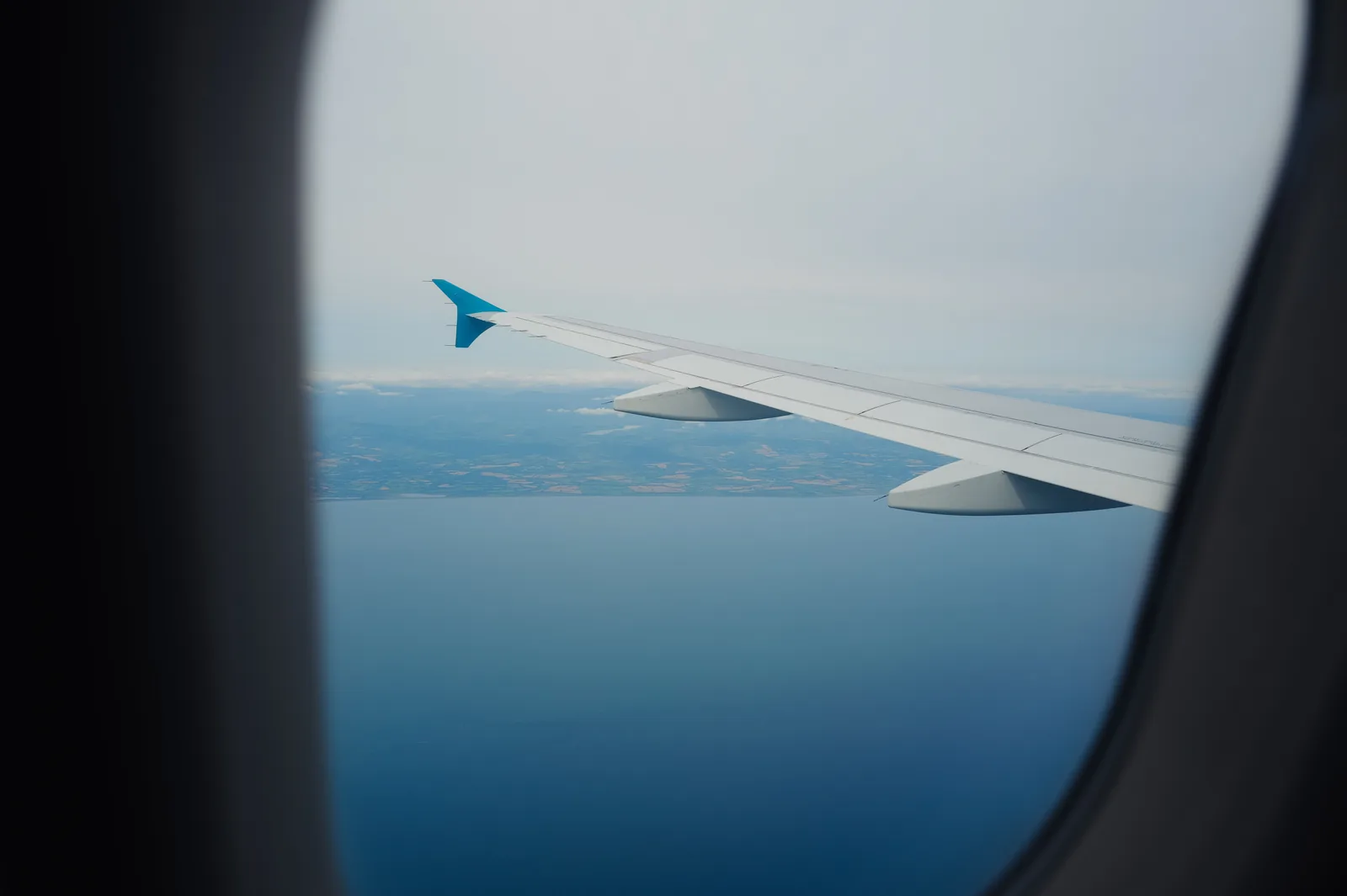 Flyvinge med blå vingespiss sett gjennom flyvinduet, over et blått hav og en flat kystlinje