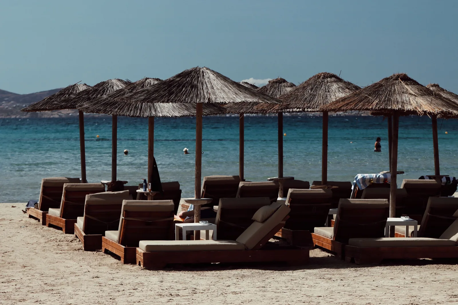 Solsenger under stråparasoller på en sandstrand ved turkist hav i Hellas