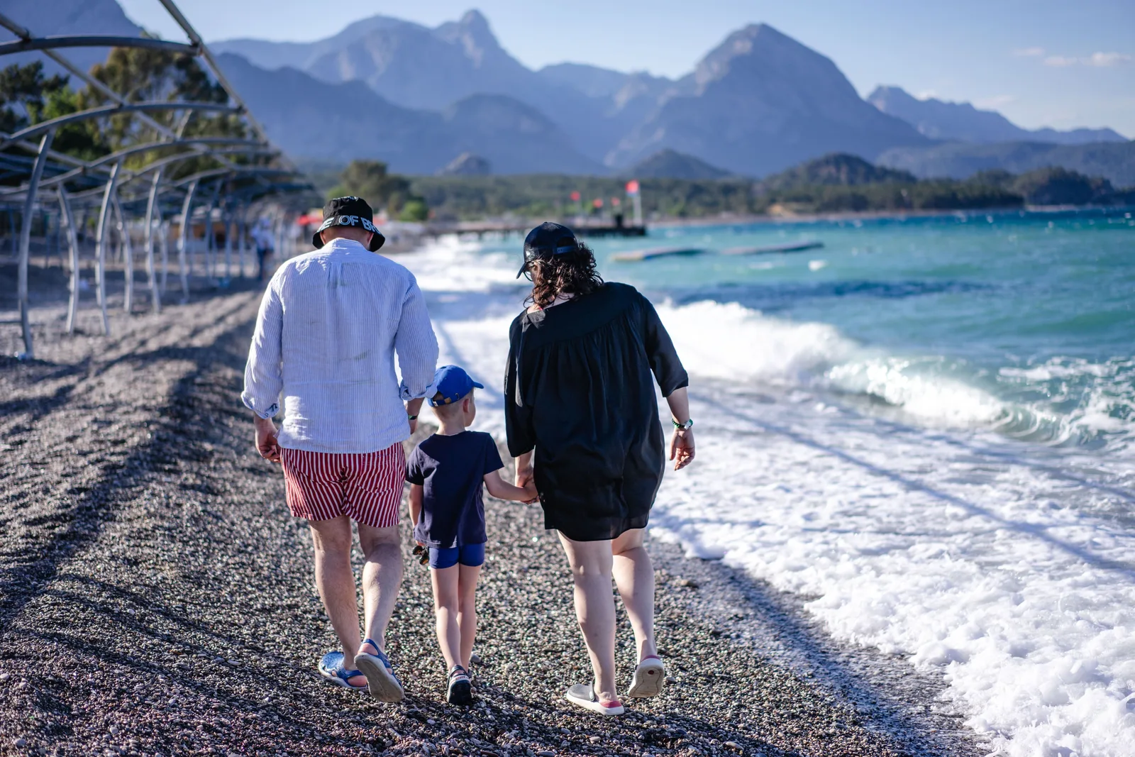 Foreldre går hånd i hånd med et barn langs en steinete strand med bølger og fjell i bakgrunnen ved Kemer i Tyrkia